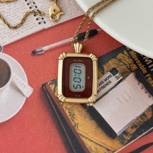 Puede incluir: Un collar dorado con un colgante de reloj rectangular. La esfera del reloj muestra n&uacute;meros digitales. El colgante tiene una esfera de color burdeos y un marco dorado. El collar descansa sobre una superficie roja con un libro y una taza de caf&eacute;.