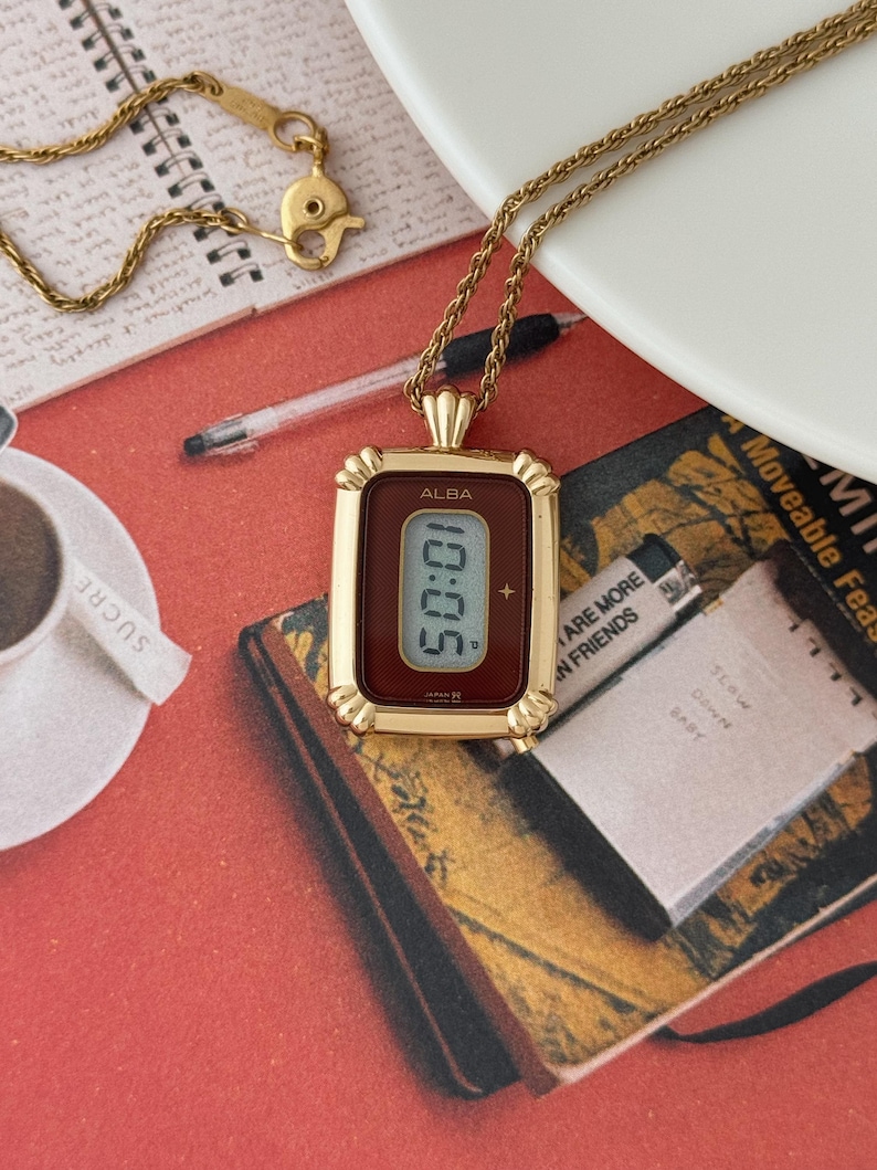 Puede incluir: Un collar dorado con un colgante de reloj rectangular. La esfera del reloj muestra n&uacute;meros digitales. El colgante tiene una esfera de color burdeos y un marco dorado. El collar descansa sobre una superficie roja con un libro y una taza de caf&eacute;.
