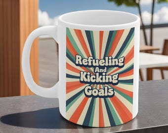 May include: White ceramic mug with a colorful design featuring the text "Refueling And Kicking Goals." The design includes a retro-style burst of orange, teal, and cream colors. The mug has a curved handle.