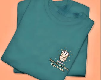 May include: Two teal crew neck sweatshirts are folded and stacked. One sweatshirt has an embroidered graphic of a coffee cup with the text "Maybe She's Born With? Maybe It's Caffeine!" in black script.