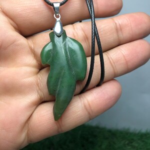 Puede incluir: Un colgante de hoja de jade verde en un collar de cordón negro. El colgante está tallado en forma de hoja con una anilla de plata.