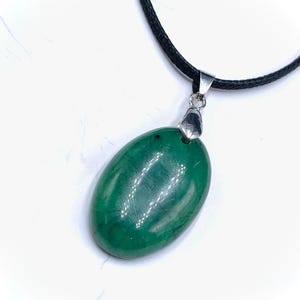 Könnte beinhalten: Eine grüne ovale Anhänger-Halskette mit schwarzem Band. Der Anhänger ist ein polierter, glänzender grüner Stein. Die Halskette hat einen silberfarbenen Metallverschluss, der den Anhänger mit dem Band verbindet.