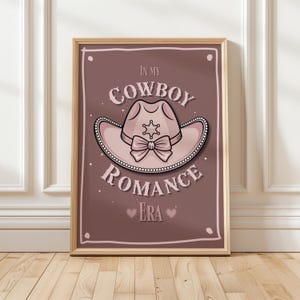 Könnte beinhalten: Gerahmter Druck mit einem rosa Cowboyhut, einem Sheriffstern und einer Schleife auf braunem Hintergrund. Der Text "IN MY COWBOY ROMANCE ERA" ist in einer stilisierten Schriftart mit Herzen dargestellt. Das Kunstwerk befindet sich in einem Holzrahmen.