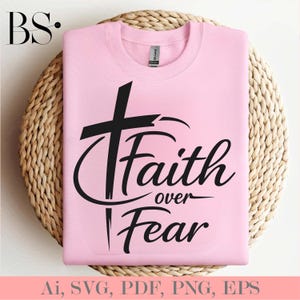 May include: A light pink sweatshirt with the text "Faith over Fear" in black script and a black cross graphic. The sweatshirt is displayed on a woven, circular placemat.