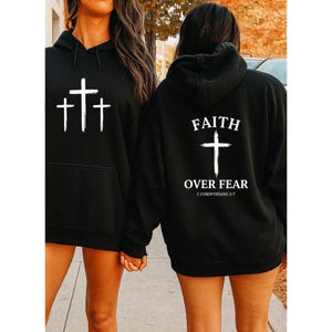 May include: Black hooded sweatshirts with white cross graphics. One sweatshirt has three crosses on the front. The other has a large cross with the words "FAITH OVER FEAR" and "2 Corinthians 5:7".