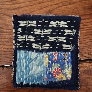 May include: Square, patchwork coaster with a dark blue border. The top section features a white dragonfly pattern on a dark blue background. The bottom sections have blue floral and abstract patterns with white stitching.