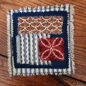May include: Square fabric coaster with a navy blue border and frayed edges. The coaster features four different fabric patterns: a cream and navy striped fabric, a red and cream floral design, a tan and red scallop pattern, and a red and white floral design.