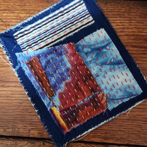 May include: A square textile art piece with a dark blue border. The piece is composed of several fabric patches in various patterns and colors, including red, blue, and white. The patches are stitched together with contrasting thread.