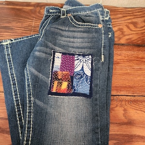 May include: Blue denim jeans with a colorful patchwork design on the back pocket. The patch features various fabric patterns in shades of purple, blue, orange, and white, stitched onto the denim with white thread. The jeans have white stitching and a classic five-pocket design.