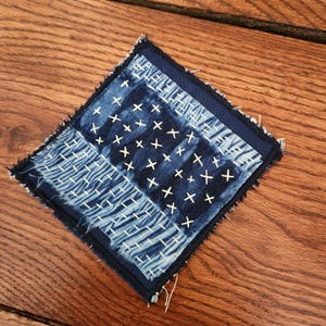 May include: Square indigo blue fabric coaster with white cross-stitch and striped patterns. The coaster has a dark blue border and frayed edges, showcasing a handcrafted aesthetic. The fabric is placed on a wooden surface.