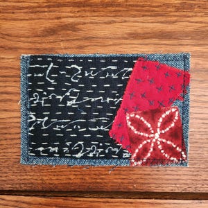 May include: A rectangular textile art piece with a denim border. The center features a dark blue fabric with white stitching and script, along with red fabric patches. One red patch has white cross stitches, and another has a white floral pattern.