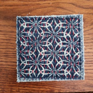 May include: A square fabric patch featuring a dark blue denim base with a repeating white geometric floral pattern. The design is accented by vertical rows of red running stitches that follow the lines of the pattern, finished with a dark blue stitched edge.