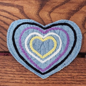 May include: A denim heart-shaped patch with embroidered concentric hearts in black, purple, white, and yellow. The patch is on a wooden surface. The design is a symbol of non-binary pride.