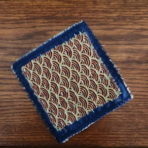 May include: A square fabric coaster featuring a central panel with a gold and dark red repeating wave pattern. The central panel is bordered by a dark blue fabric frame with frayed edges and visible white stitching along the perimeter.