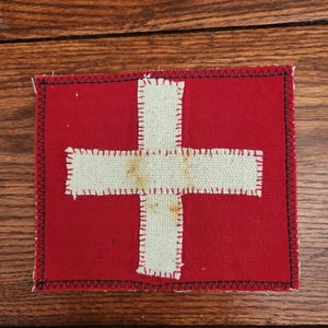 May include: A rectangular red patch with a white cross in the center, resembling a flag. The cross is made of a textured, off-white material and is stitched onto the red background. The edges are finished with black stitching.