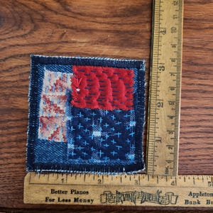 May include: A square fabric patch featuring a patchwork design of dark blue denim, a red textured section, and a light patterned fabric with blue stitching. A wooden ruler shows the patch measures approximately 3 inches by 3 inches.