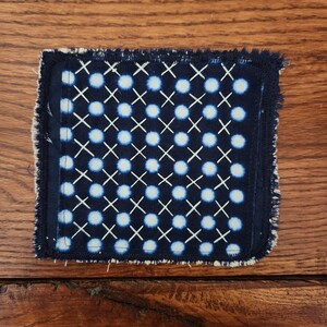 May include: Square navy blue and white patterned textile with a repeating design of light blue circles and white crosses. The edges are frayed, giving it a rustic appearance. The textile is placed on a wooden surface.
