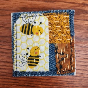 May include: A square fabric patch featuring a patchwork design with denim borders. The center panel displays two yellow and black bees on a white honeycomb background. Adjacent panels include mustard yellow and brown fabric with visible yellow stitching.
