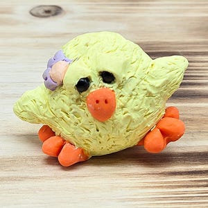 May include: A handcrafted yellow chick figurine with orange feet and beak, and black eyes. The chick has a textured surface and a small purple flower on its head. This decorative item is perfect for Easter.