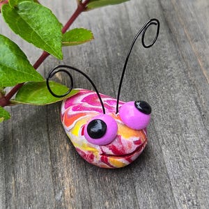 May include: A colorful, handcrafted stone bug with pink, yellow, and red swirls. It has pink eyes with black pupils and black wire antennae. Green leaves and a red stem are in the background.