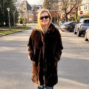May include: A woman wearing a long, dark brown faux fur coat and blue jeans stands on a paved road. She is wearing sunglasses and smiling. The coat has a large collar and pockets. The background includes trees, buildings, and parked cars.