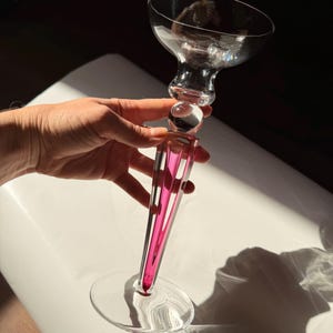 May include: A clear glass champagne coupe with a pink and clear stem. The stem has a pink stripe design. The coupe is held up by a hand against a white surface. The glass has a round base.