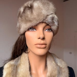 May include: A vintage fur hat and fur collar in shades of brown and tan. The hat is a rounded shape, and the collar is draped around the mannequin's shoulders. The fur appears soft and luxurious, suggesting a high-end fashion item.