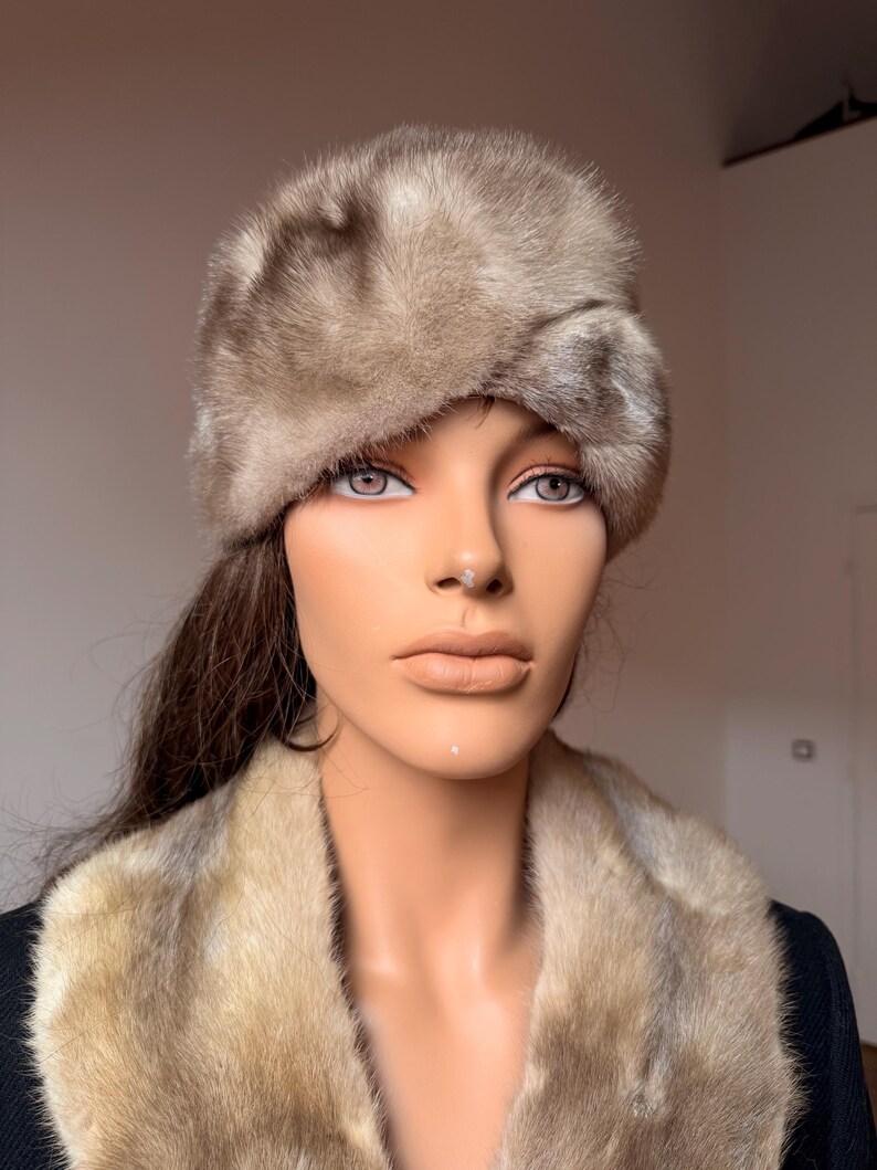 May include: A vintage fur hat and fur collar in shades of brown and tan. The hat is a rounded shape, and the collar is draped around the mannequin's shoulders. The fur appears soft and luxurious, suggesting a high-end fashion item.