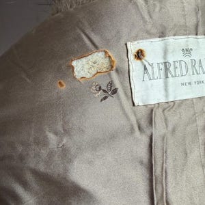 Pode incluir: Close-up do forro interior de um casaco vintage. O forro &eacute; de cor bege suave com uma pequena rosa bordada e a etiqueta "ALFRED RA NEW YORK". H&aacute; uma rasgadura no forro.
