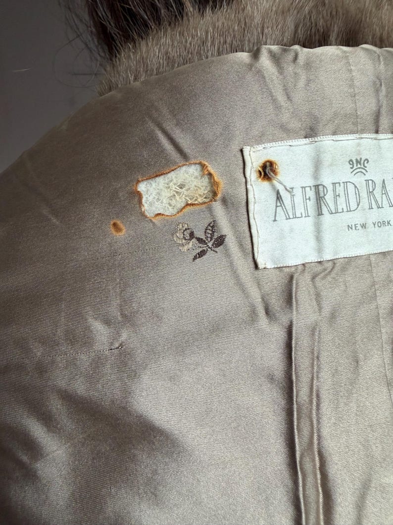 Pode incluir: Close-up do forro interior de um casaco vintage. O forro &eacute; de cor bege suave com uma pequena rosa bordada e a etiqueta "ALFRED RA NEW YORK". H&aacute; uma rasgadura no forro.