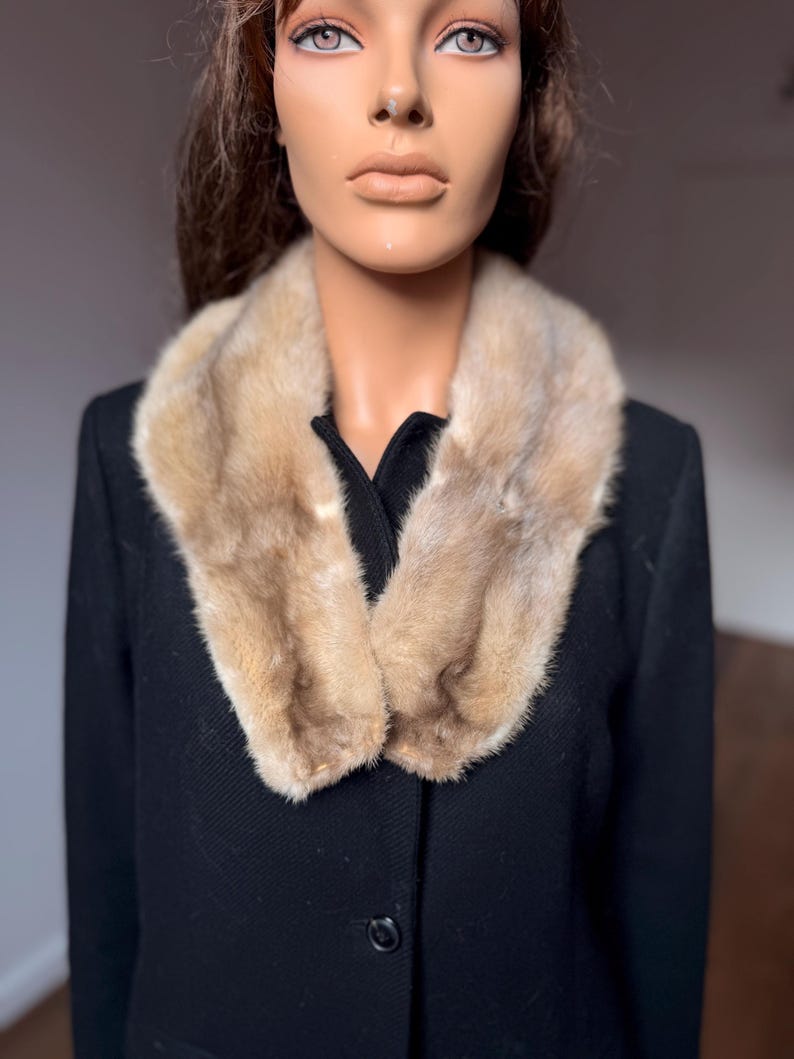 May include: A black coat with a light brown fur collar. The coat has a classic design with a button closure. The fur collar adds a touch of luxury and warmth to the garment. The coat is displayed on a mannequin.