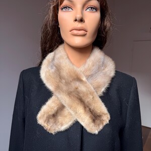 May include: A light brown fur scarf is draped over a black coat. The fur scarf is made of soft, light brown fur. The coat is a solid black color. The mannequin is wearing the scarf and coat.