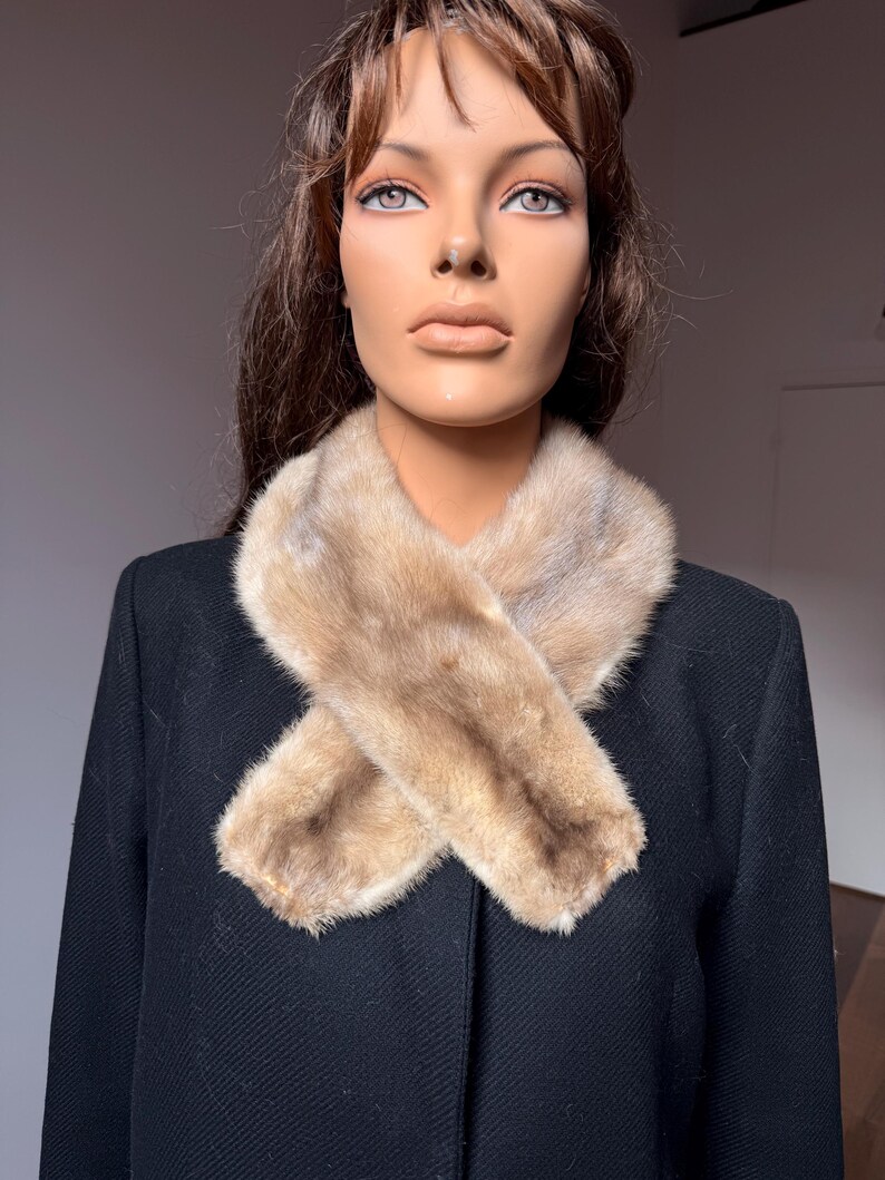 May include: A light brown fur scarf is draped over a black coat. The fur scarf is made of soft, light brown fur. The coat is a solid black color. The mannequin is wearing the scarf and coat.