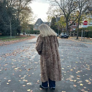 Pu&ograve; includere: Un lungo cappotto in finta pelliccia marrone con un motivo strutturato. Il cappotto &egrave; indossato sopra pantaloni blu scuro e scarpe nere. La persona &egrave; in piedi su una strada bagnata e coperta di foglie con alberi ed edifici sullo sfondo.