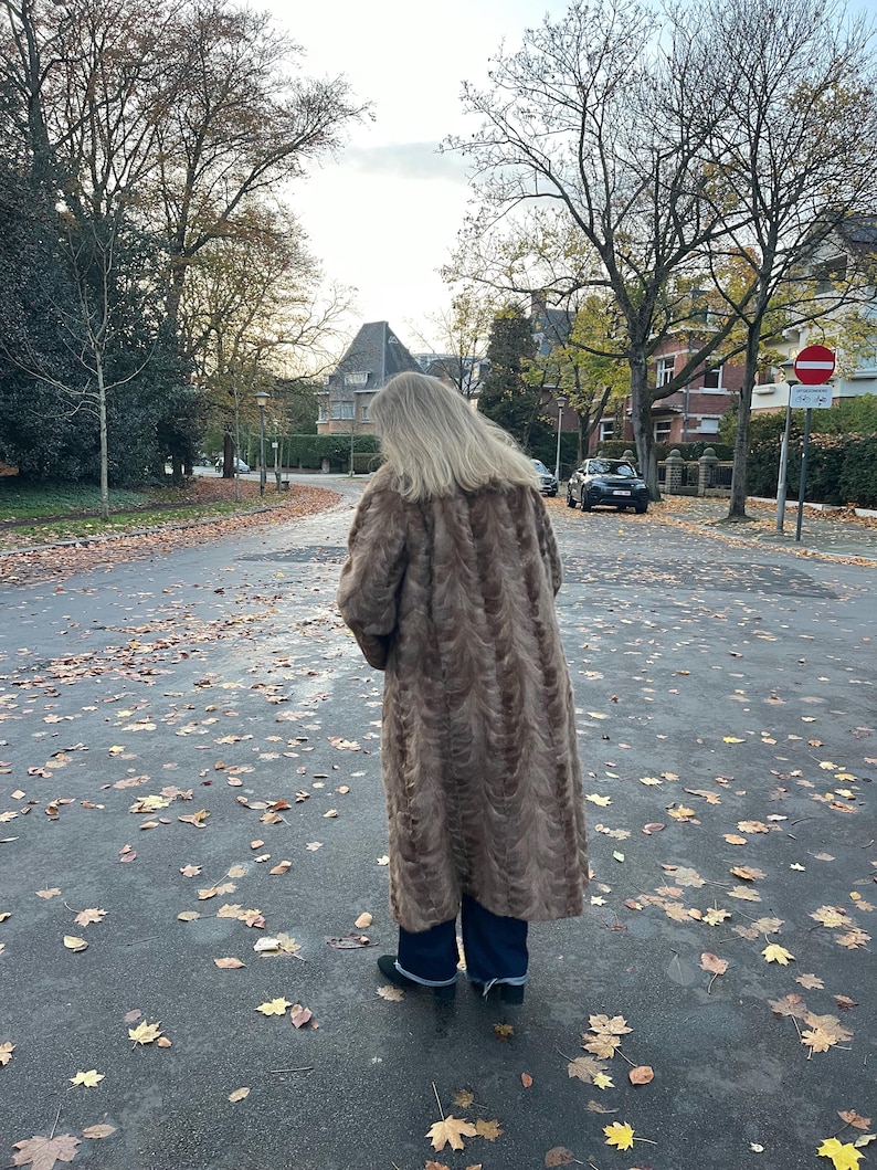 Pu&ograve; includere: Un lungo cappotto in finta pelliccia marrone con un motivo strutturato. Il cappotto &egrave; indossato sopra pantaloni blu scuro e scarpe nere. La persona &egrave; in piedi su una strada bagnata e coperta di foglie con alberi ed edifici sullo sfondo.