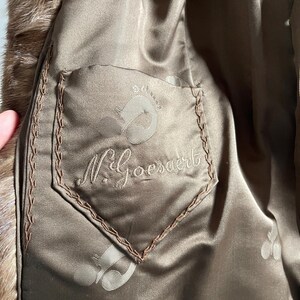 Pu&ograve; includere: Primo piano dell'interno di un cappotto di pelliccia marrone. La fodera &egrave; un materiale simile al raso con una tasca con il nome "N. Goesaert" ricamato e un logo. I bordi della tasca sono cuciti con filo marrone.