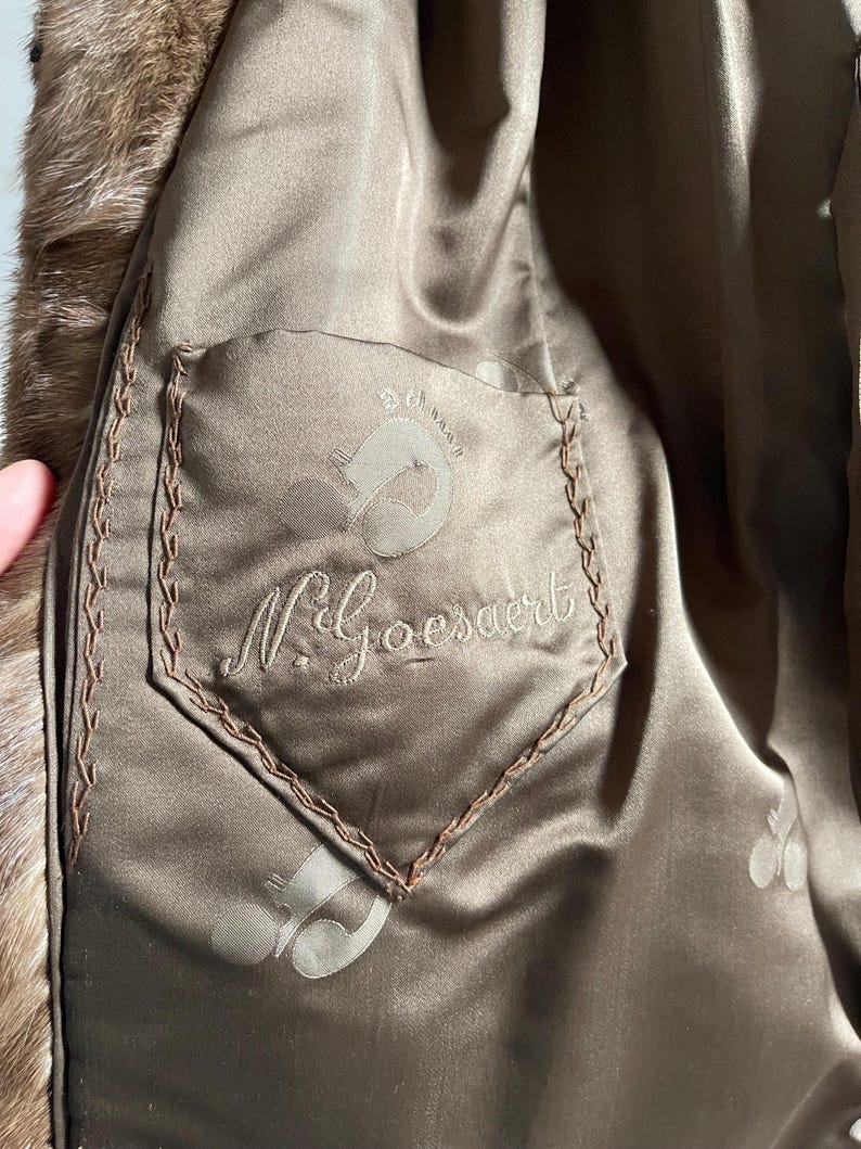 Pu&ograve; includere: Primo piano dell'interno di un cappotto di pelliccia marrone. La fodera &egrave; un materiale simile al raso con una tasca con il nome "N. Goesaert" ricamato e un logo. I bordi della tasca sono cuciti con filo marrone.