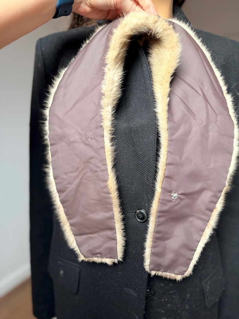 May include: A brown and beige fur collar with a dark brown lining. The collar is draped over a black coat, showcasing the contrast between the textures and colors. A small decorative button is visible on the coat.