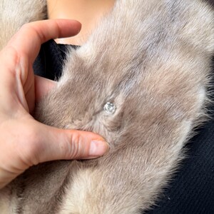 May include: A light brown fur collar with a sparkling, round, clear crystal clasp. The fur is soft and fluffy, with a slight sheen. The collar is worn over a black garment.