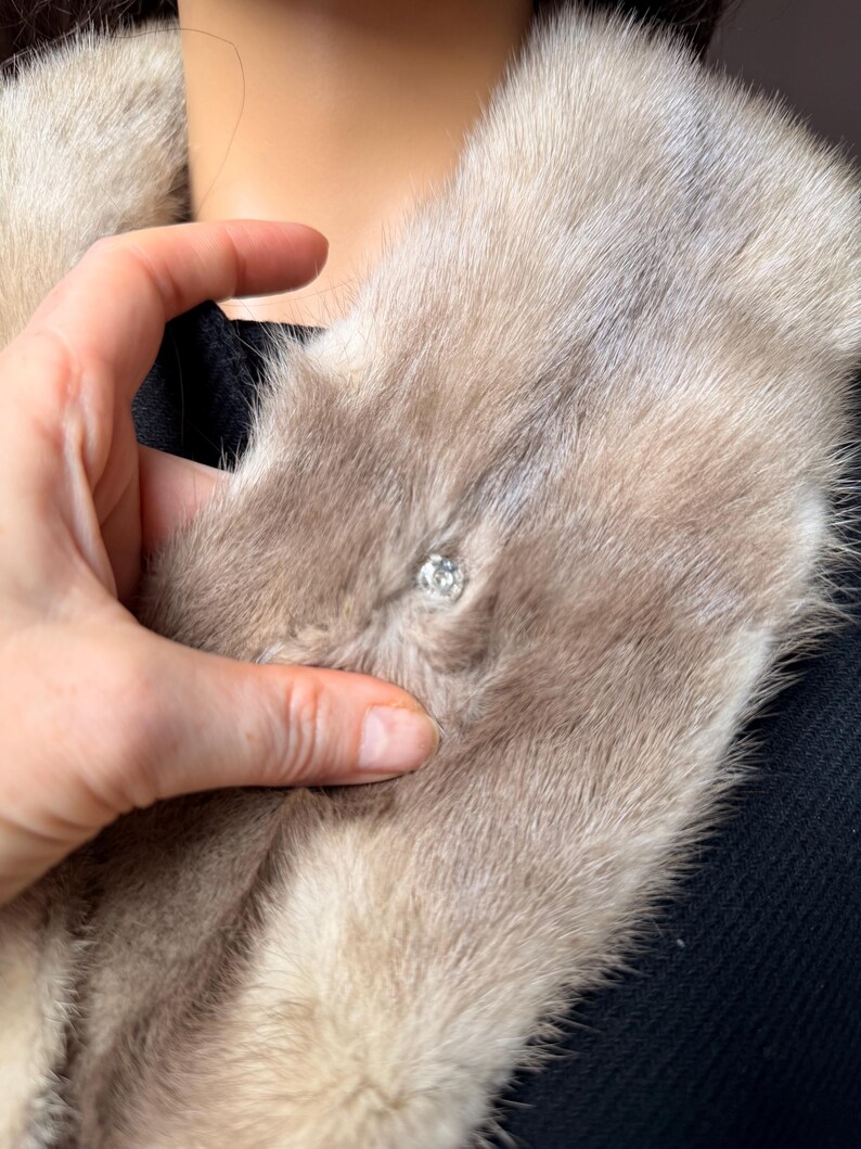 May include: A light brown fur collar with a sparkling, round, clear crystal clasp. The fur is soft and fluffy, with a slight sheen. The collar is worn over a black garment.
