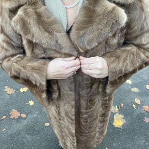 Pu&ograve; includere: Un lungo cappotto di pelliccia marrone con un ampio colletto e chiusura con bottoni. Il cappotto sembra essere realizzato in un materiale morbido e soffice. Il cappotto &egrave; indossato sopra una sciarpa colorata e un top nero.