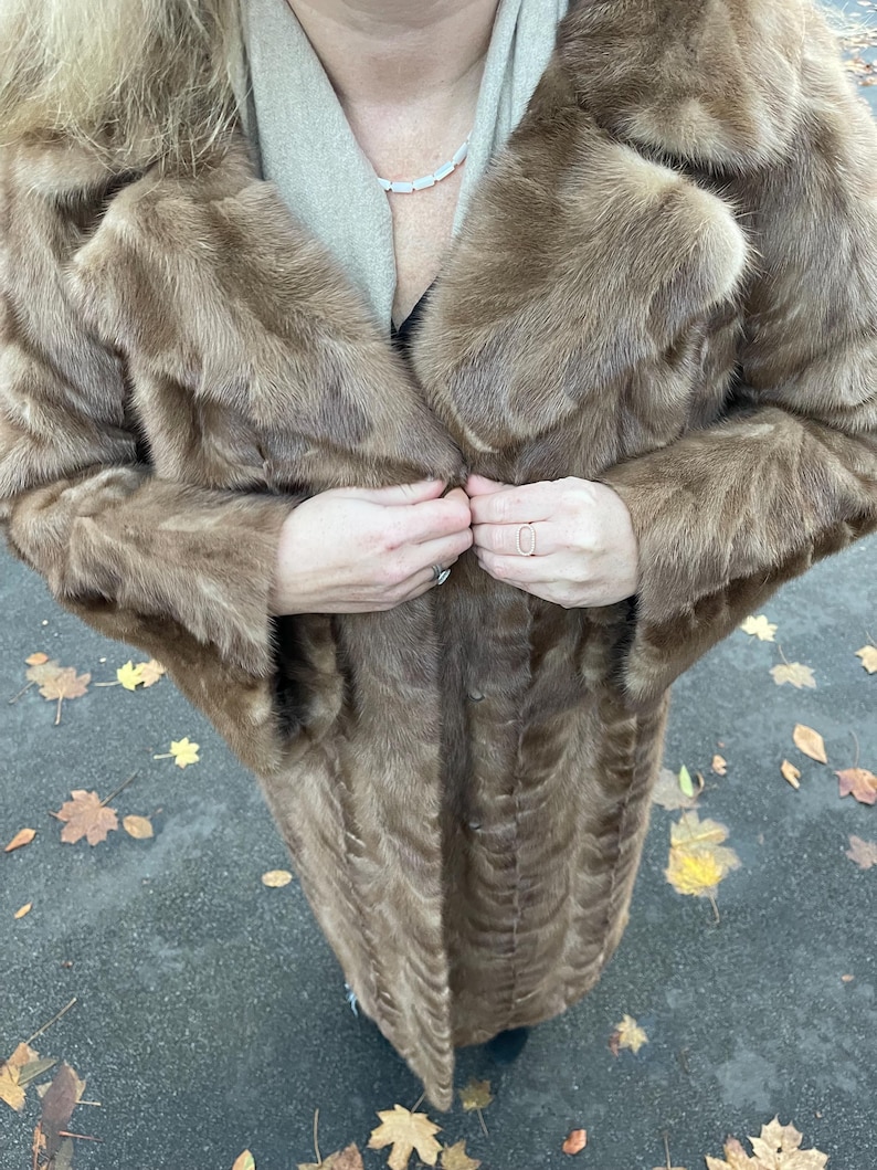 Pu&ograve; includere: Un lungo cappotto di pelliccia marrone con un ampio colletto e chiusura con bottoni. Il cappotto sembra essere realizzato in un materiale morbido e soffice. Il cappotto &egrave; indossato sopra una sciarpa colorata e un top nero.