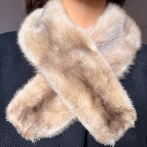 May include: A light brown fur scarf, with a crossed design. The scarf is made of soft, fluffy fur and is a neutral color. The scarf is displayed on a black background.