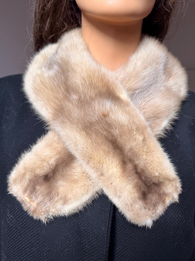 May include: A light brown fur scarf, with a crossed design. The scarf is made of soft, fluffy fur and is a neutral color. The scarf is displayed on a black background.