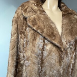 Pu&ograve; includere: Un cappotto di pelliccia marrone con colletto classico e chiusura con bottoni. Il cappotto ha un aspetto morbido e strutturato e un colore marrone medio. Il cappotto &egrave; esposto su un manichino.