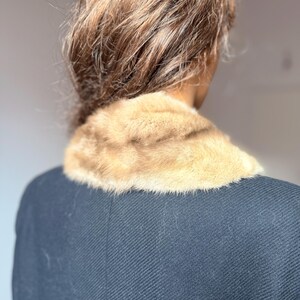 May include: A close-up view of a vintage black coat with a light brown fur collar. The fur collar is soft and plush, contrasting with the dark, textured fabric of the coat. The coat's design is classic and elegant.
