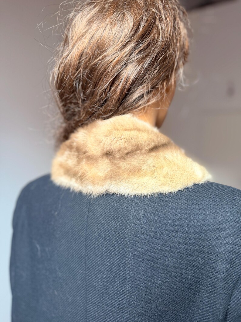 May include: A close-up view of a vintage black coat with a light brown fur collar. The fur collar is soft and plush, contrasting with the dark, textured fabric of the coat. The coat's design is classic and elegant.