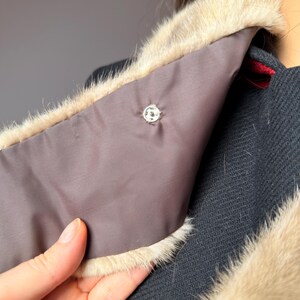 May include: Close-up of a black coat with a light brown fur collar. The collar has a dark brown lining and a decorative silver button. The coat's texture is visible, and a hand is holding the collar.