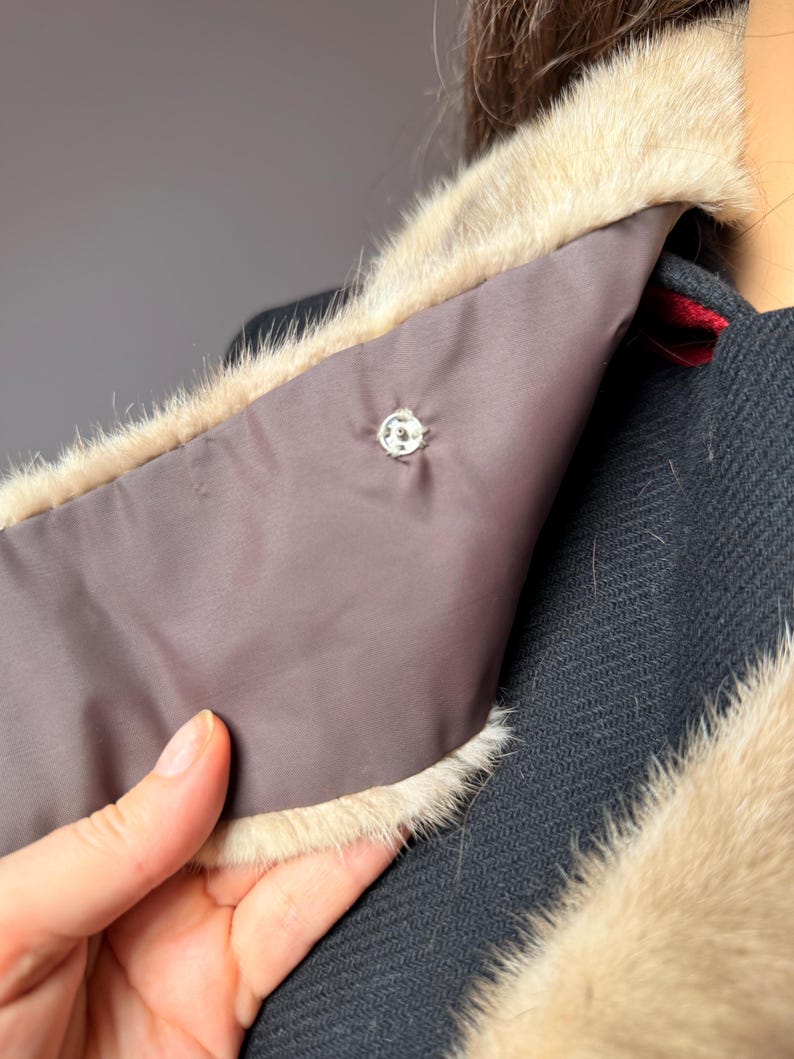 May include: Close-up of a black coat with a light brown fur collar. The collar has a dark brown lining and a decorative silver button. The coat's texture is visible, and a hand is holding the collar.