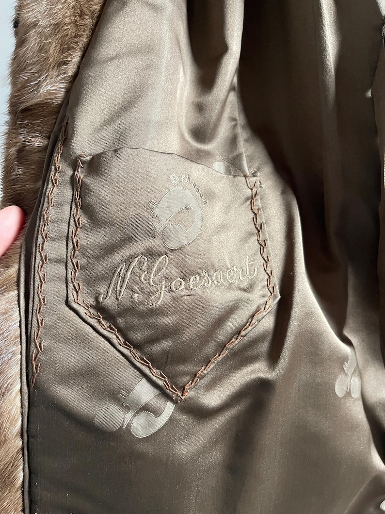 Pu&ograve; includere: Primo piano dell'interno di un cappotto di pelliccia marrone. La fodera &egrave; in materiale satinato con una tasca con il nome ricamato "N. Goesaert" e un logo. I bordi della tasca sono cuciti con filo marrone.